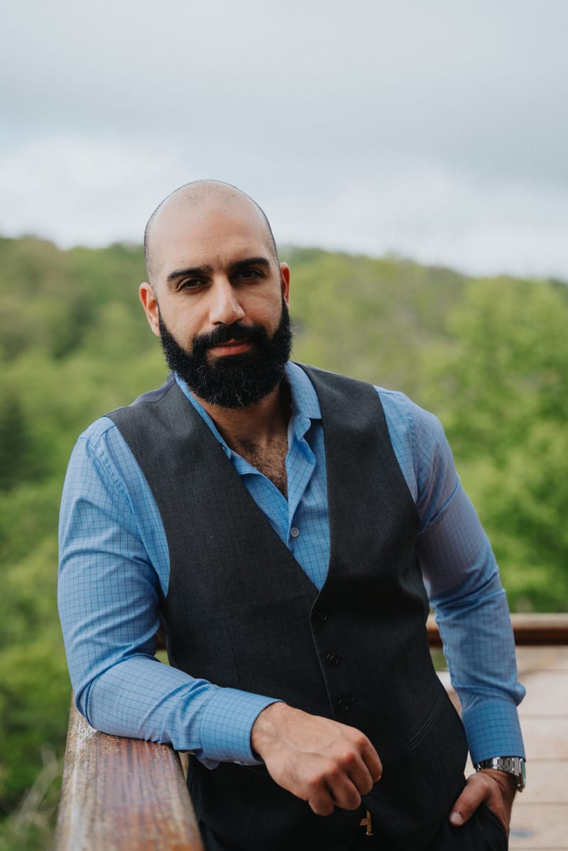 Portrait of Ayman Al-Abdullah in a vest leaning on a wooden railing with green hills behind
