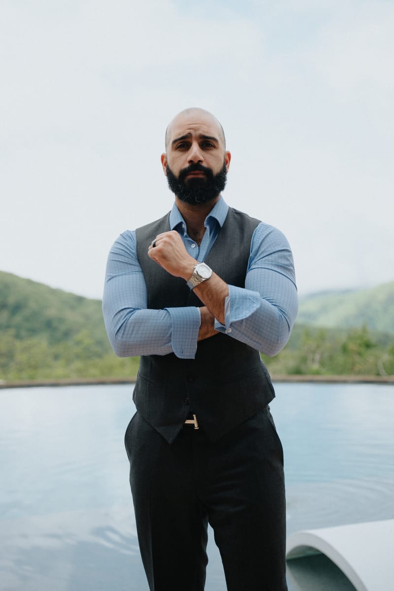 Portrait of Ayman Al-Abdullah in a vest adjusting his watch, standing by a pool with hills behind